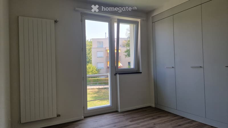 Empty room with wooden flooring, white walls, built-in closet, and large window with a view of the outside area.