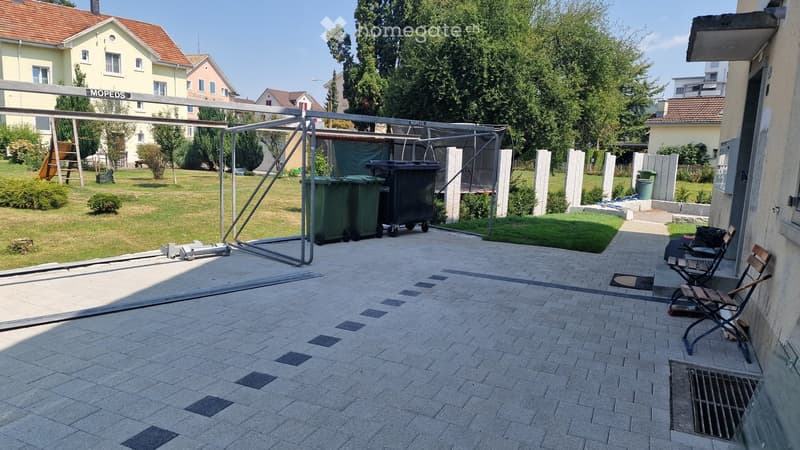 Outdoor patio area with brick pavement, two folding chairs, green trash bins, and a grassy lawn with trees and houses in the background.