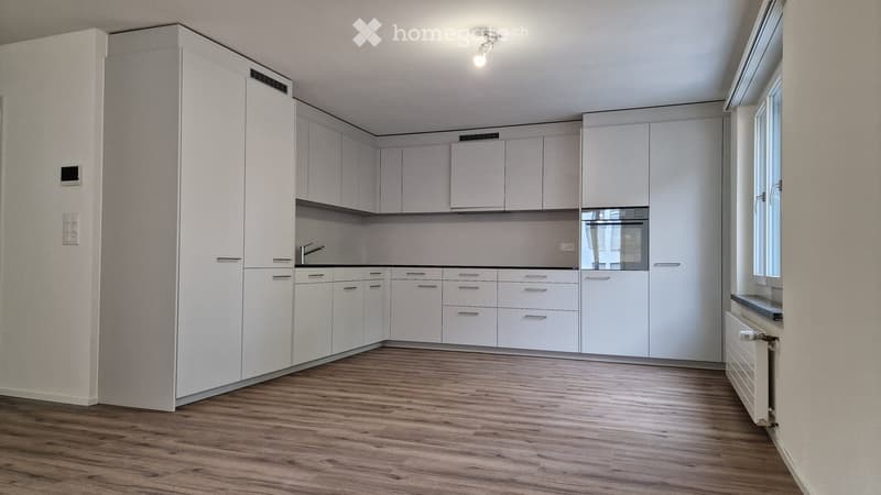 Empty kitchen with white cabinets, wooden floor, and large windows. No appliances or furniture are present.
