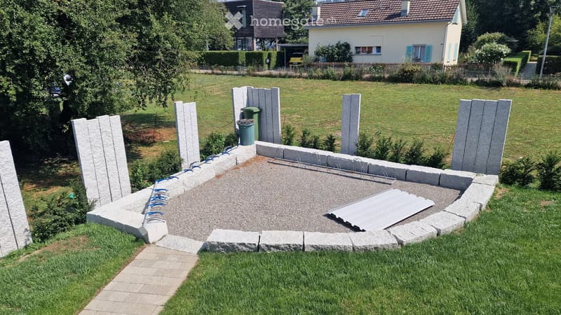 Garden area with a raised concrete platform surrounded by stone pillars, a green trash bin, and a metal cover on the ground.
