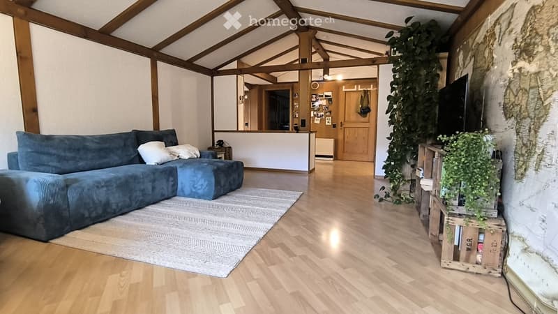 Modern living room with exposed wooden beams, hardwood flooring, a blue sectional sofa, a coffee table, a television, and a large leafy plant in the corner.