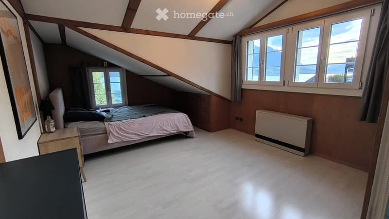 Cozy attic bedroom with slanted wooden ceiling, large windows offering a scenic view, a neatly made bed, a nightstand, and a radiator.