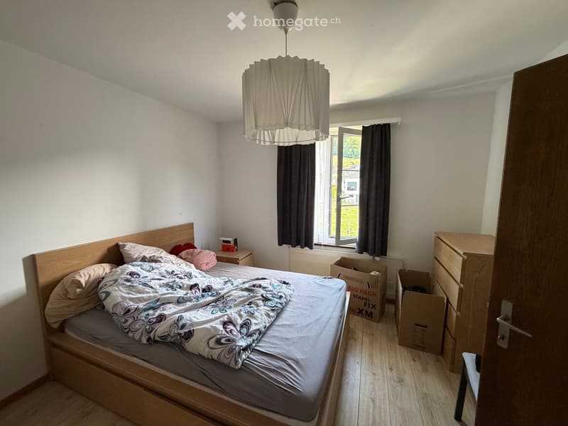 Bedroom with a wooden bed, nightstand, lamp, chest of drawers, and a stool. The room has wooden flooring and white walls.
