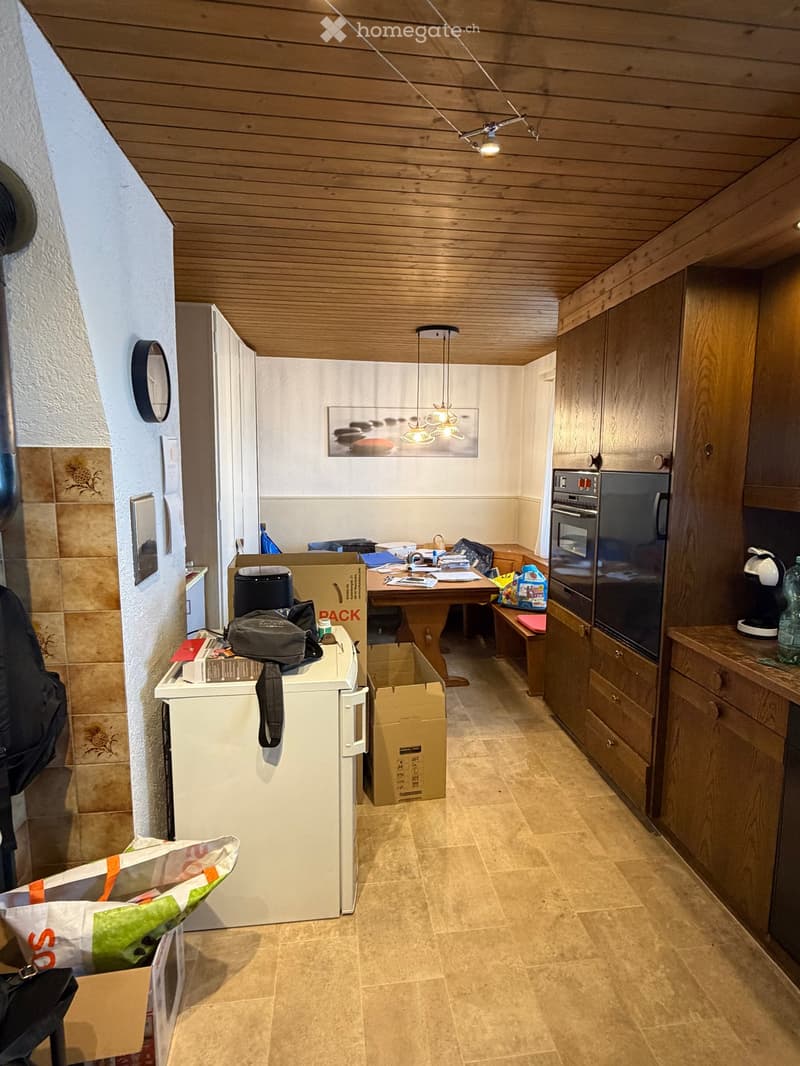 A furnished room with a refrigerator, oven, table, chairs, cabinets, and a lamp. The room has wooden ceilings and tiled floors.