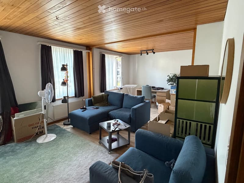 Living room with a blue sectional sofa, coffee table, fan, and a small table with chairs. Wooden ceiling and large windows with curtains.