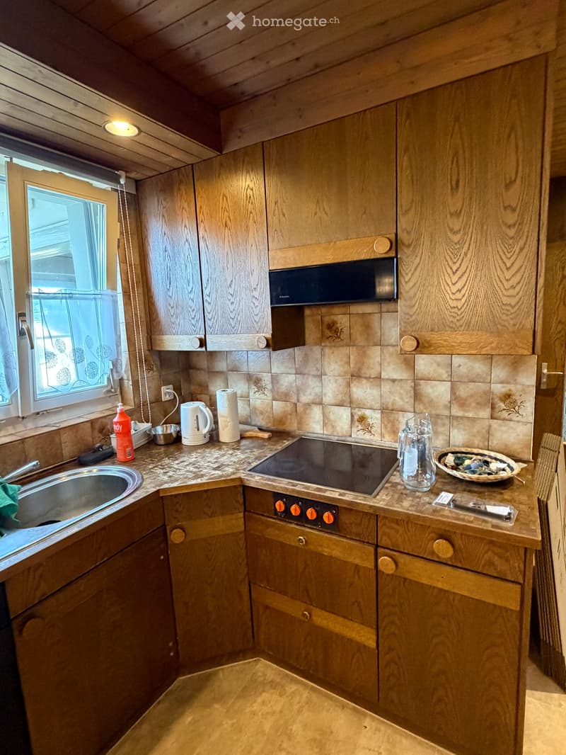 The kitchen features wooden cabinets, a tiled backsplash, and a countertop with various appliances and items, including a sink, electric kettle, oven, and microwave.
