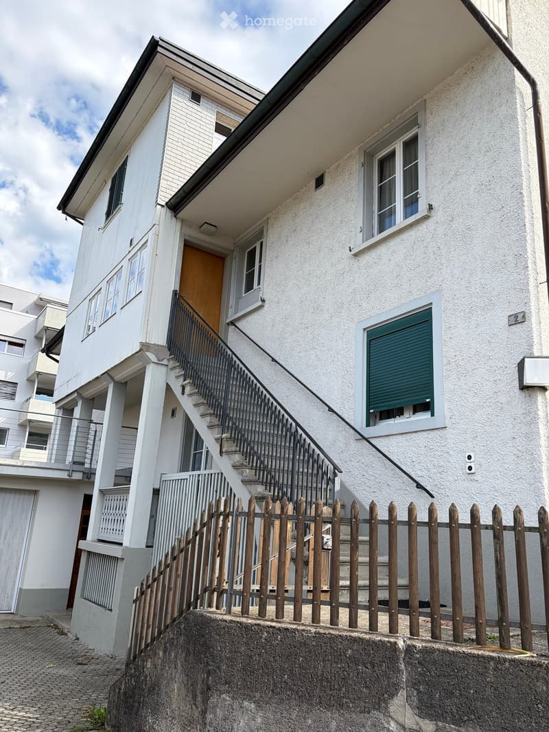 Two-story house with white exterior, raised ground floor, staircase leading to entrance, wooden fence, and windows with shutters.