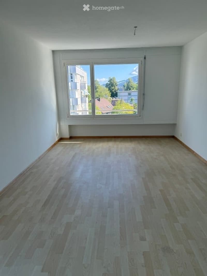 Empty room with wooden flooring and large window offering a view of buildings and greenery outside.