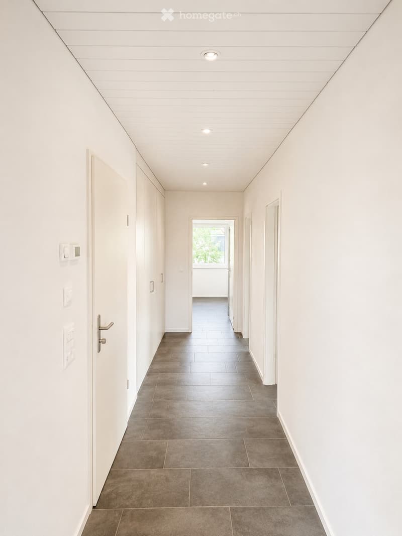 A modern, minimalist hallway with white walls, recessed ceiling lighting, and a tiled floor. Features include a door on the left and an open doorway leading to another room at the end.