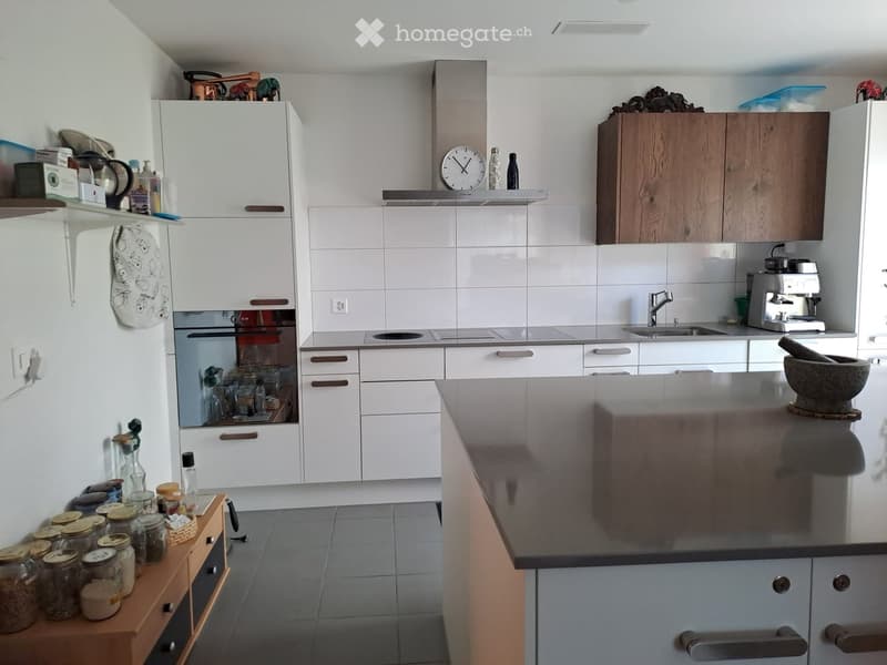 Modern kitchen with white cabinets, stainless steel appliances, and a central island. Features include a refrigerator, oven, sink, faucet, coffee maker, clock, and various jars and bottles on the counter.