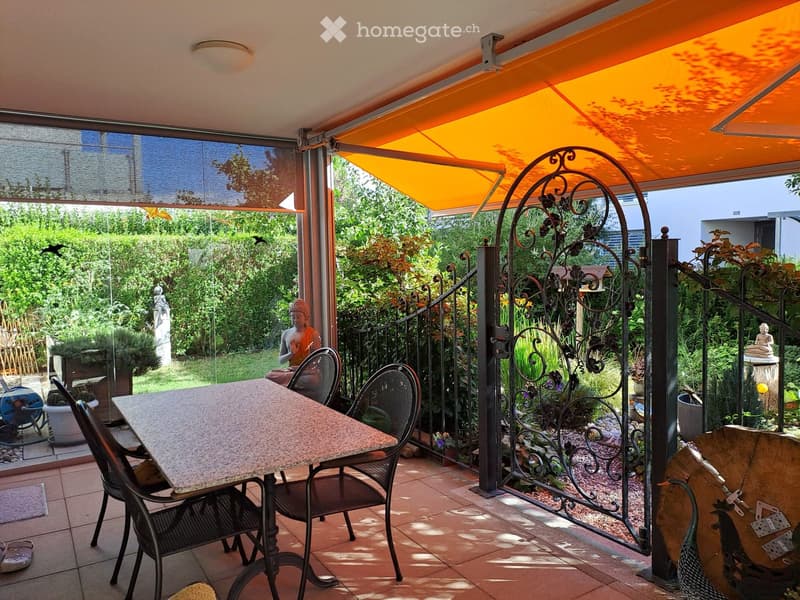 A covered patio with a table and chairs, overlooking a lush garden with statues and decorative elements.