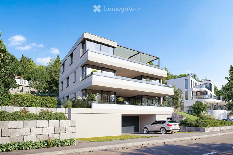 Modern two-story house with white exterior, large windows, and a balcony on the upper level. A car is parked in the driveway, and the house is surrounded by greenery and a stone wall.