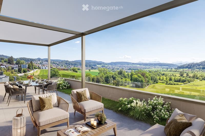 A spacious balcony with a panoramic view of a green landscape and distant mountains. It features a seating area with wicker chairs and a small table, along with a dining set for four, surrounded by potted plants and flowers.