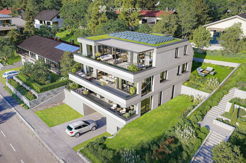 A modern two-story house with a green roof, large windows, and multiple outdoor seating areas. The house features a spacious terrace on the upper level and a well-maintained garden with a staircase leading to the entrance.