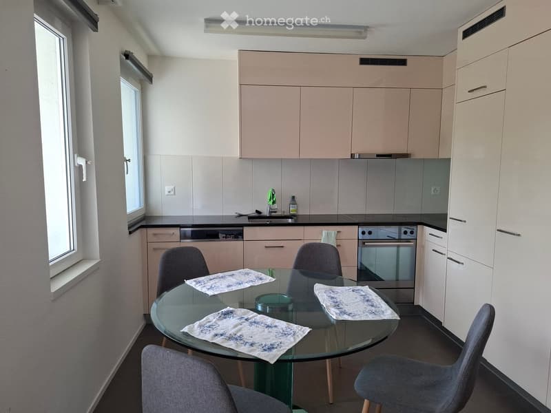 Modern kitchen with light-colored cabinets, stainless steel appliances, a glass dining table with four chairs, and a window providing natural light.
