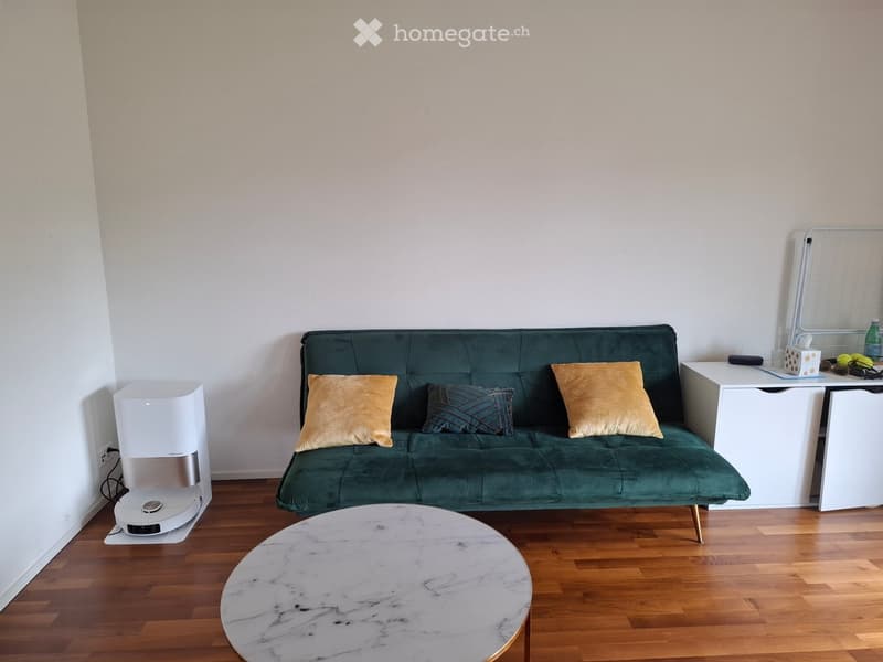 A modern living room with a green velvet sofa, decorative pillows, a round marble coffee table, a white speaker, and a small table with a mirror and various items on top.