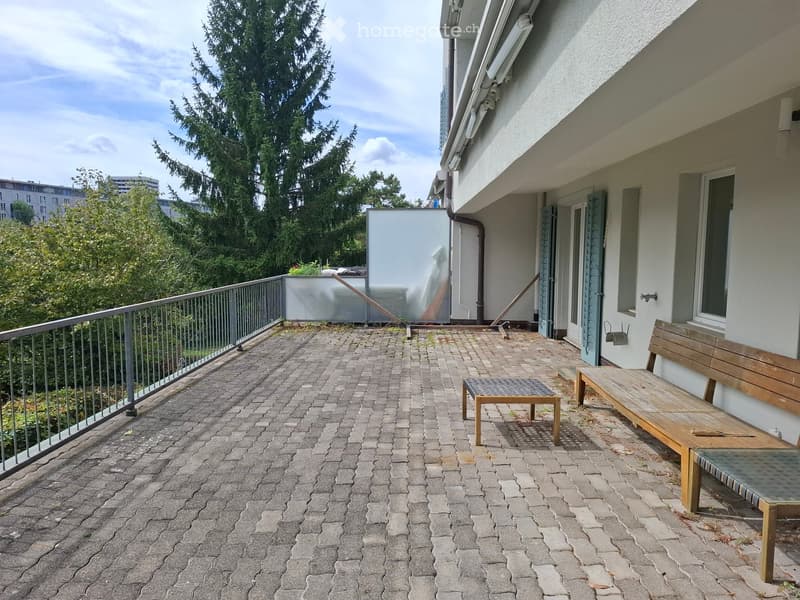Balcony with brick pavement, metal railing, wooden bench, small table, and a view of greenery and buildings.