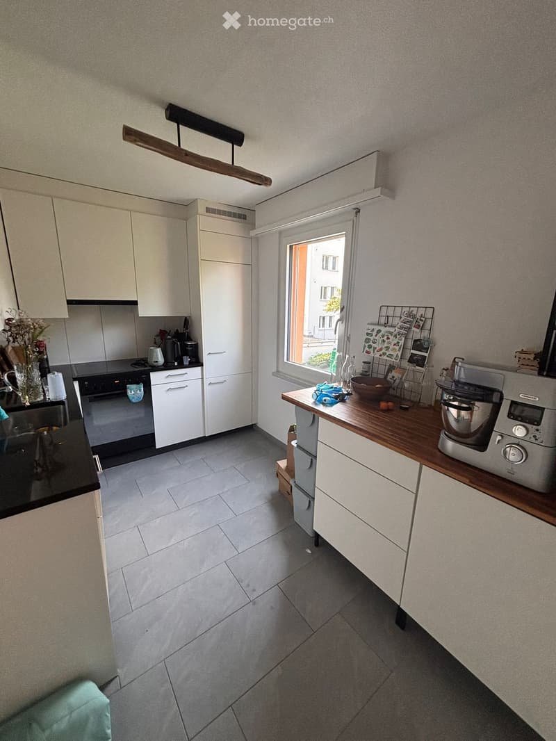 Modern kitchen with white cabinets, grey tiles, and a wooden island. Appliances include a refrigerator, oven, microwave, and blender. A window offers a view of the street.