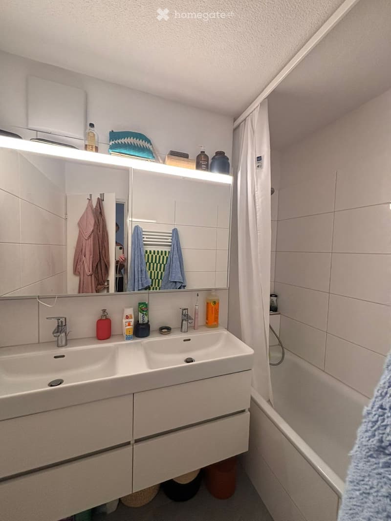 Modern bathroom with a double sink, large mirror with built-in lighting, and various toiletries on the counter. Towels and a bathrobe are visible.