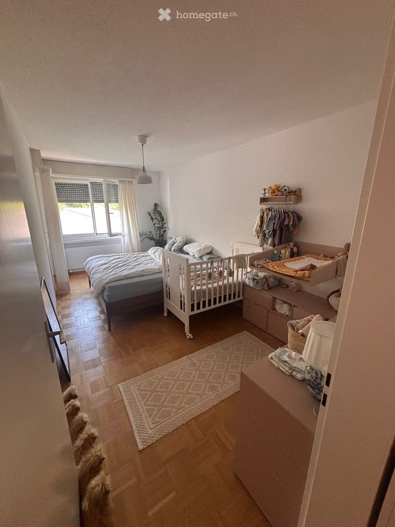 Brightly lit bedroom with wooden flooring, a double bed, a crib, a changing table, a wardrobe, and a small plant near the window.