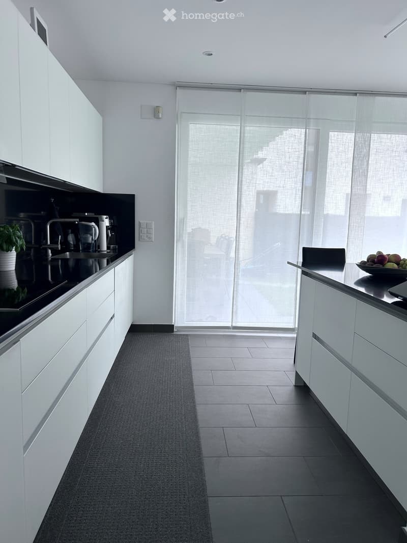 Modern kitchen with white cabinets, black countertops, and a large window with sheer curtains. Includes a sink, faucet, coffee maker, blender, bowl of fruit, and a small dining area with a table and chair.