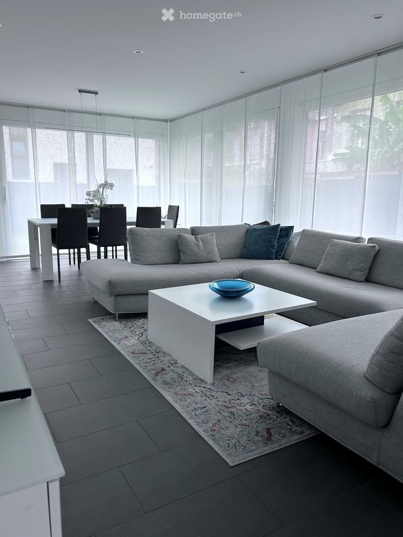 Modern living room with a grey sectional sofa, white coffee table, dining area with chairs, and large windows with sheer curtains.