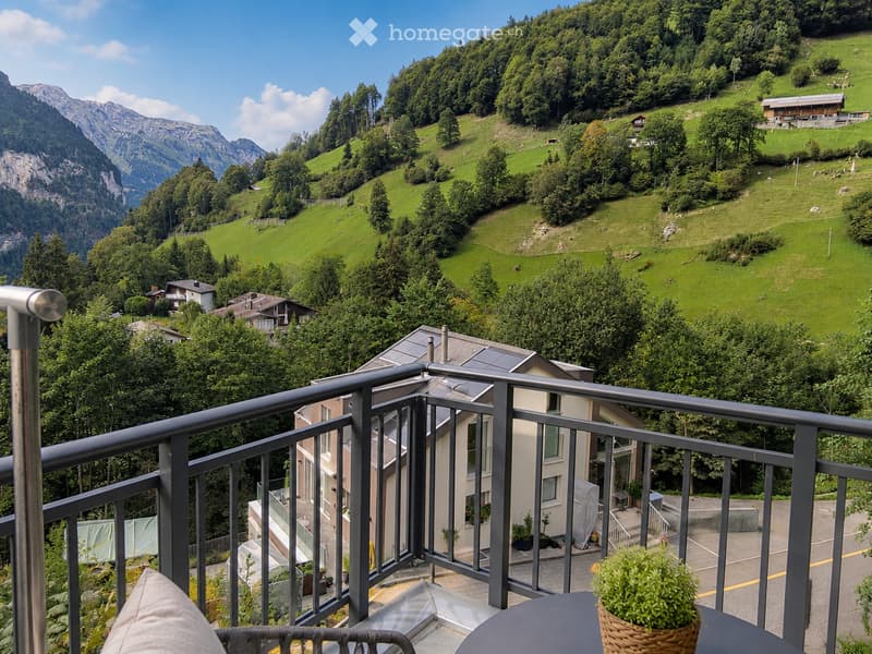 Balcony overlooking a lush green valley with rolling hills, scattered houses, and distant mountains under a clear blue sky.