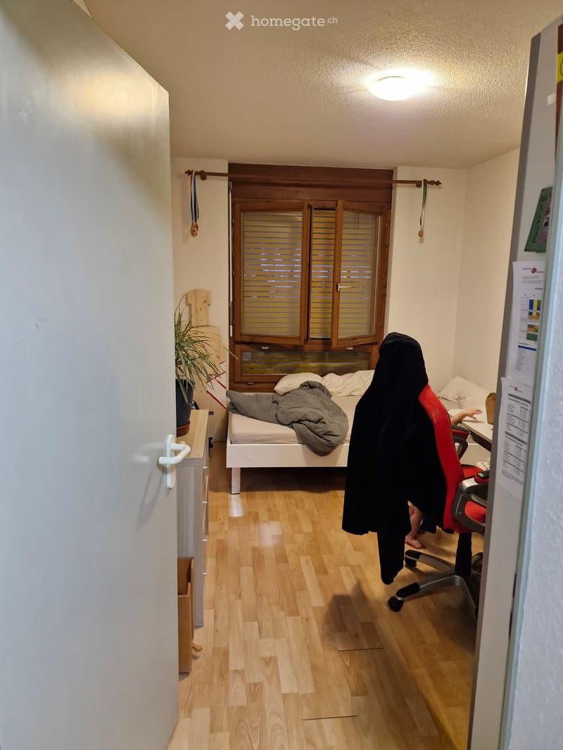 A small bedroom with a single bed, a chair, a table, and a plant near the window. The room has wooden flooring and a slatted window with blinds.