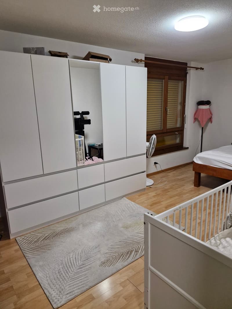 Modern bedroom with wooden flooring, large white wardrobe with mirrored doors, a bed with white bedding, a fan near the window, and a baby gate in the foreground.