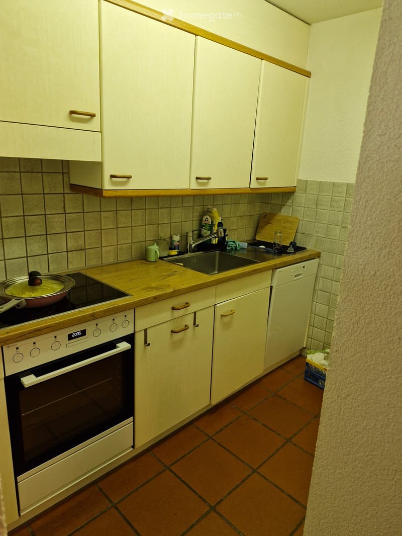 A small kitchen with white cabinets, wooden countertops, and a tiled floor. Features an oven, dishwasher, sink, and various kitchen items on the counter.