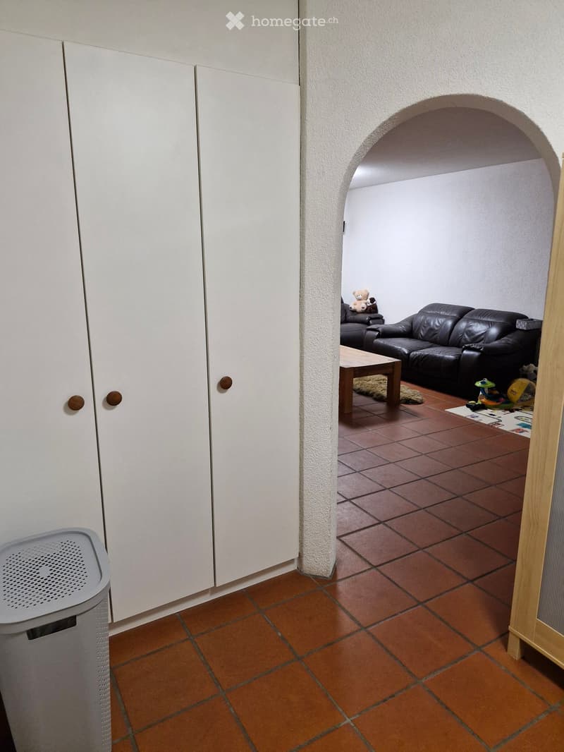 A hallway with terracotta tiled flooring, white walls, and an arched opening leading to a living area with a black leather sofa, wooden coffee table, and toys.