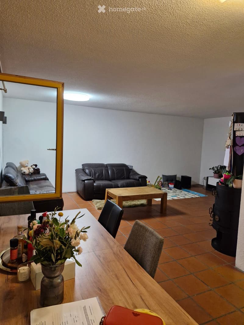 A well-lit living and dining area with a black leather sofa, wooden coffee table, dining table with chairs, and a vase of flowers on the table. The room features tiled flooring and a large mirror on the wall.