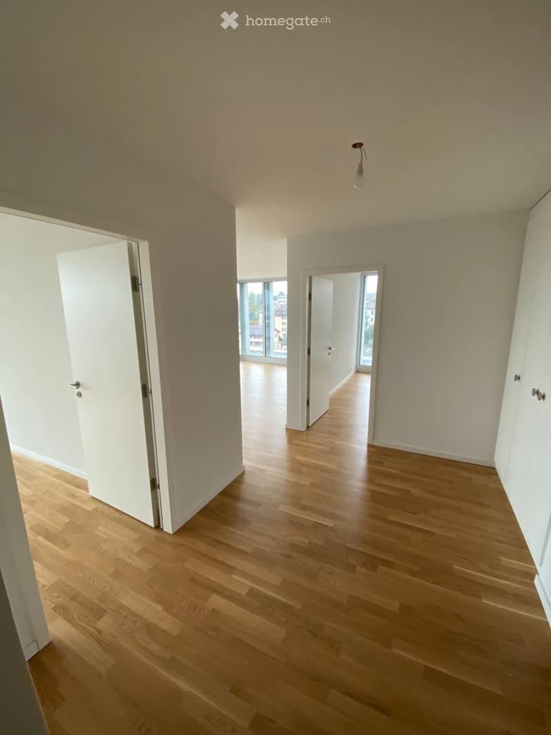 Empty room with light wooden flooring, white walls, and a single ceiling light fixture. The room has two open doors leading to other spaces.