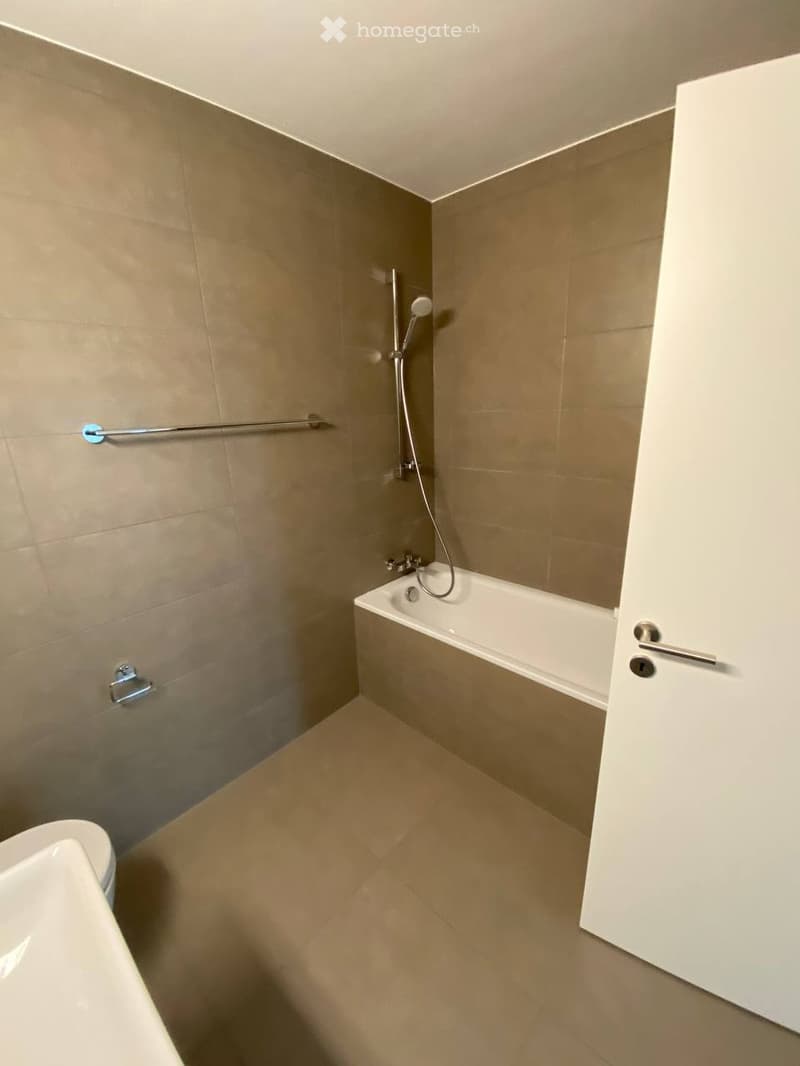 Modern bathroom with a bathtub, toilet, and a showerhead, featuring beige tiled walls and a handrail for safety.
