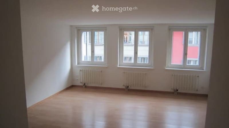 Empty room with wooden flooring, white walls, and three windows with white frames. Radiators are present under each window.