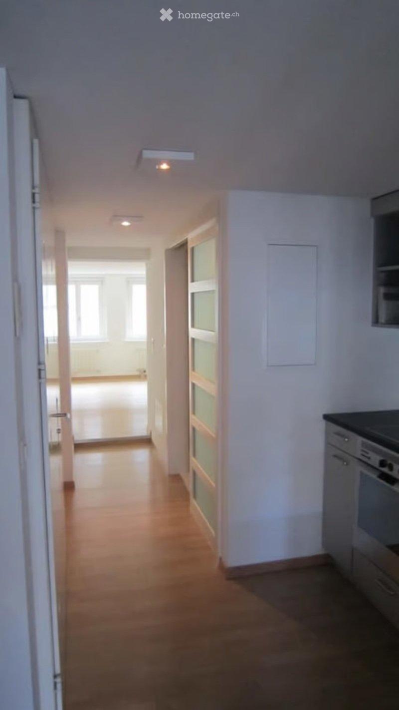 Empty hallway with wooden flooring, white walls, and recessed lighting. Doors lead to adjacent rooms, including a kitchen area visible on the right.
