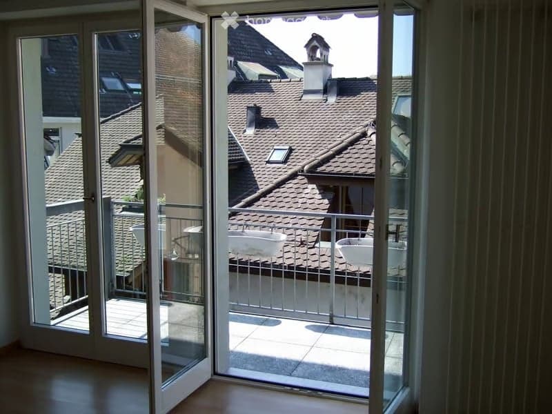 Balcony with metal railing, overlooking neighboring rooftops and chimneys. The balcony is accessible through large glass doors from the interior.