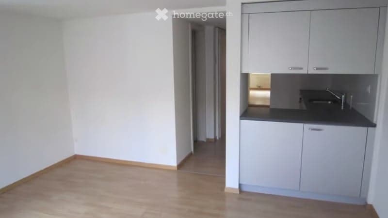 Empty room with light wooden flooring and white walls, featuring an open doorway leading to another room and a small kitchen area with cabinets and a sink.