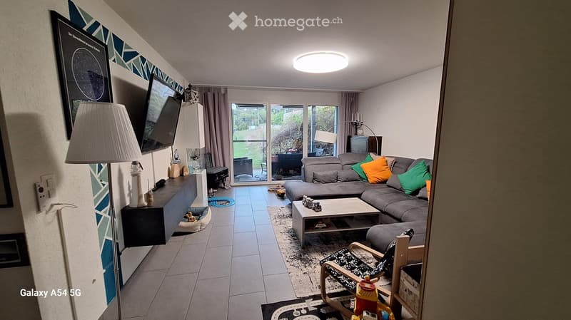Modern living room with a grey sectional sofa, colorful throw pillows, a white coffee table, a wooden chair, a floor lamp, a mounted TV screen, and a pet bed near the sliding glass doors.