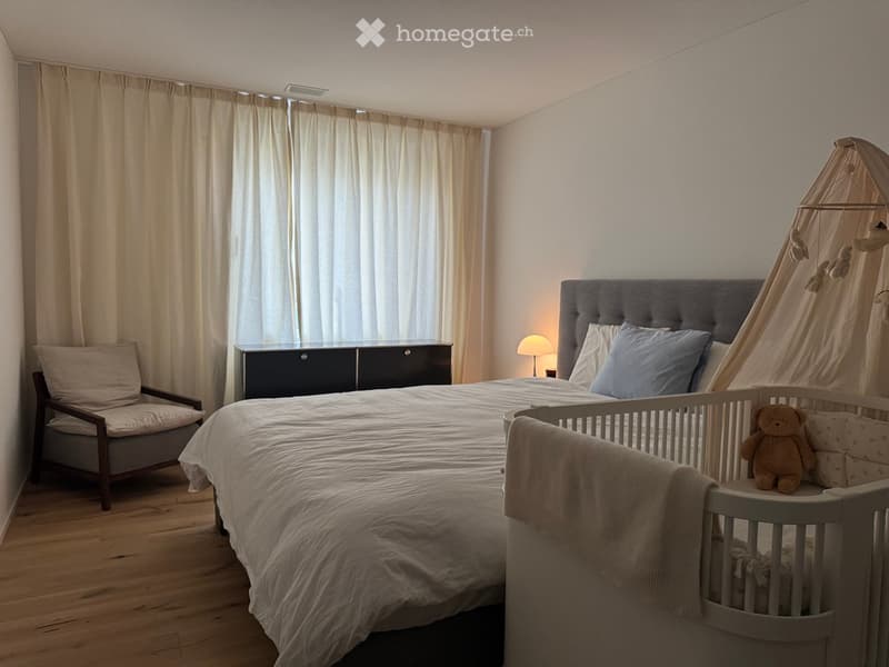 A well-lit bedroom with a large bed, a wooden chair, a nightstand with a lamp, a crib with a teddy bear, and a hanging mobile above the crib.