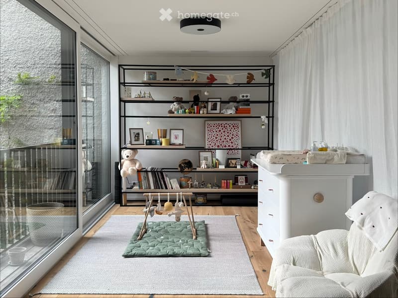 A well-lit, modern nursery with wooden flooring, large windows, a shelf filled with toys and books, a changing table, a rocking chair, and a play mat.