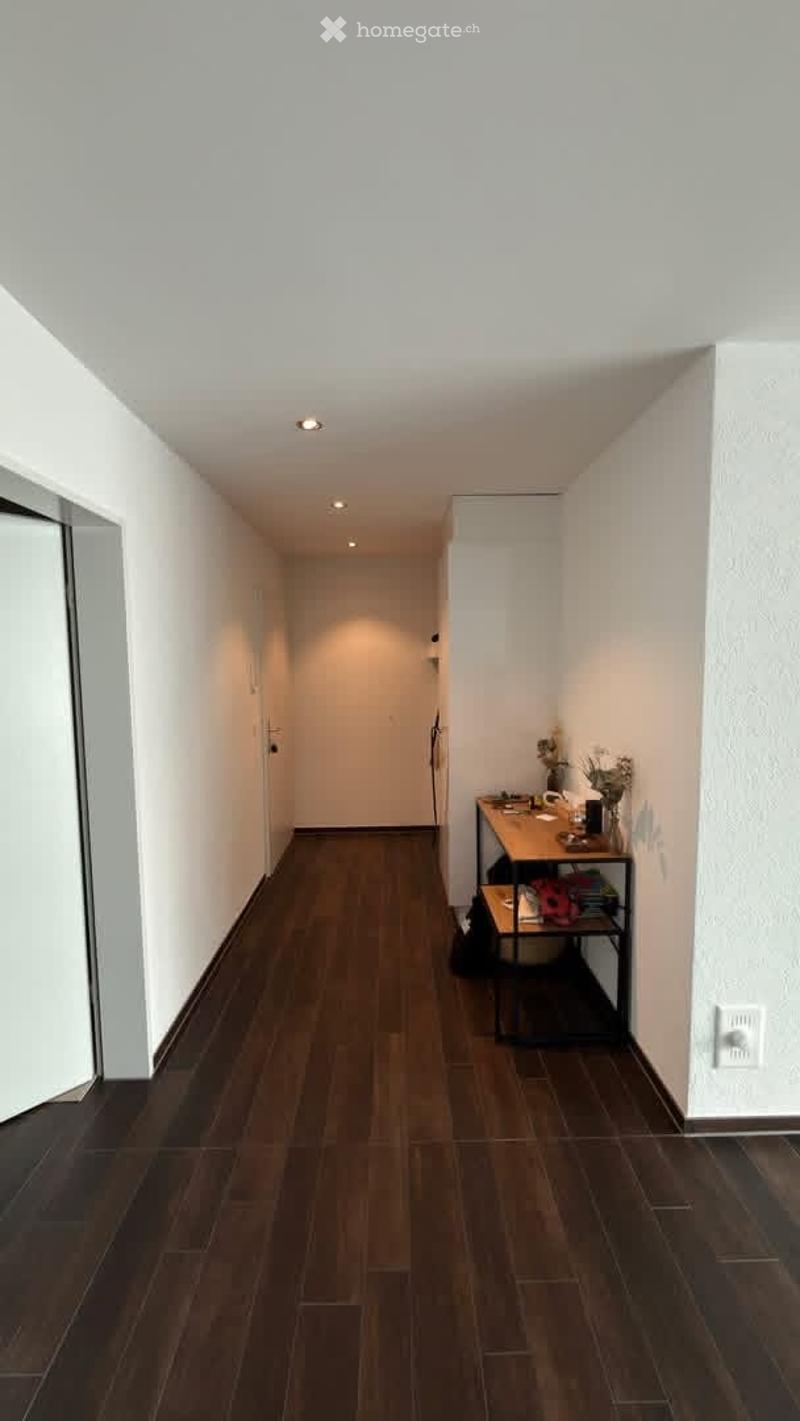 A modern hallway with wooden flooring, white walls, and recessed lighting. A small table with a vase and a shelf with various items is placed against the wall.