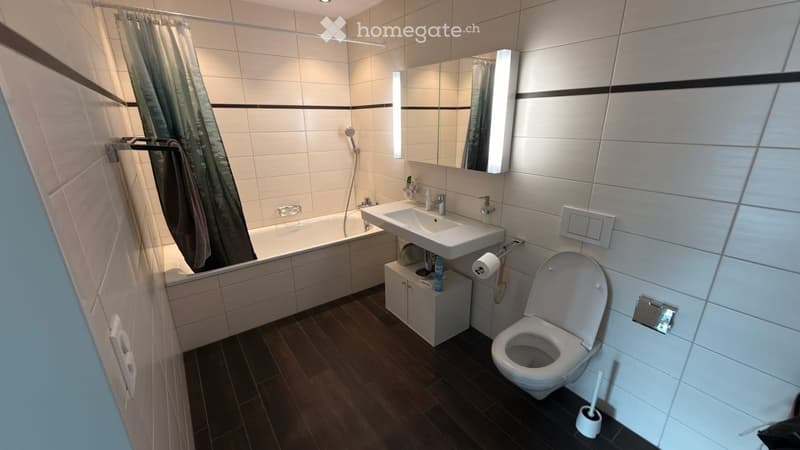 Modern bathroom with a bathtub, sink, toilet, mirror, and a shower curtain. The floor is dark wood, and the walls are tiled with white tiles and black accents.