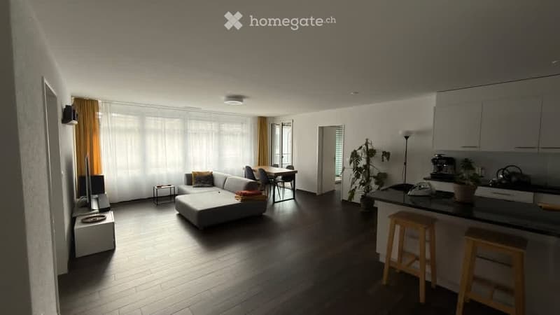 Open-plan living and dining area with a grey sectional sofa, coffee table, dining table with chairs, kitchen island with stools, and a potted plant. The space features large windows with sheer curtains and wooden flooring.