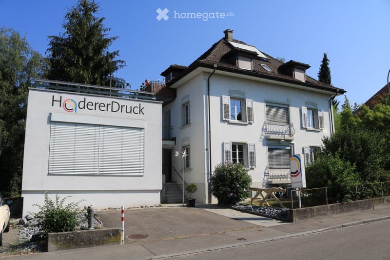 Two-story white building with a brown roof, multiple windows, and a sign reading "HödererDruck" on the side.