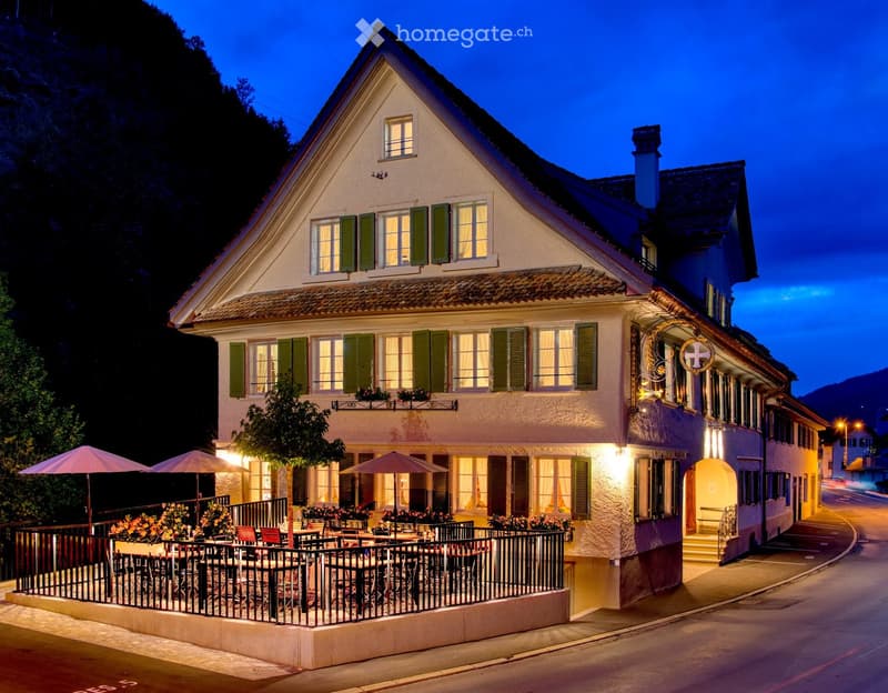 Two-story traditional house with green shutters, illuminated windows, and a small outdoor seating area with tables and chairs under umbrellas.