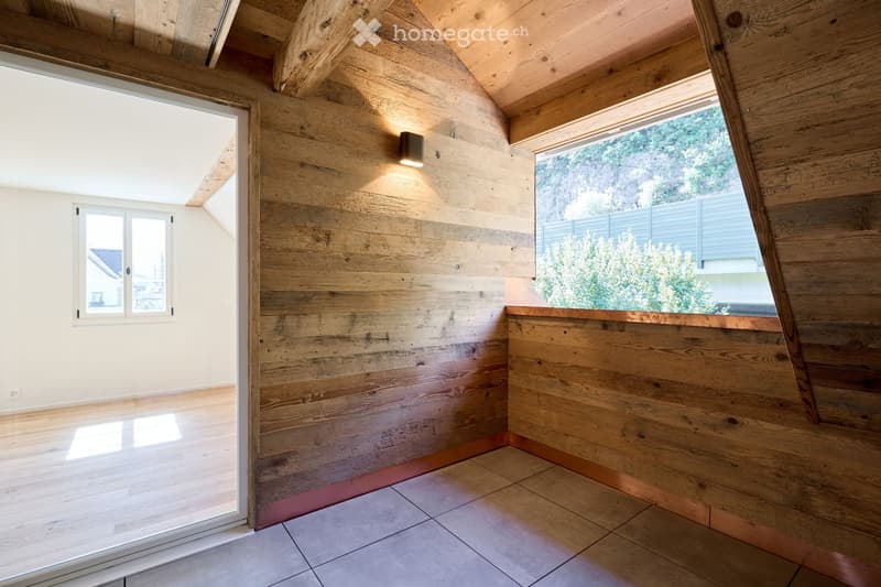 An unfurnished room with wooden walls, a slanted wooden ceiling, and large windows offering a view of greenery outside. The floor is tiled, and the room features built-in lighting and a sliding glass door.