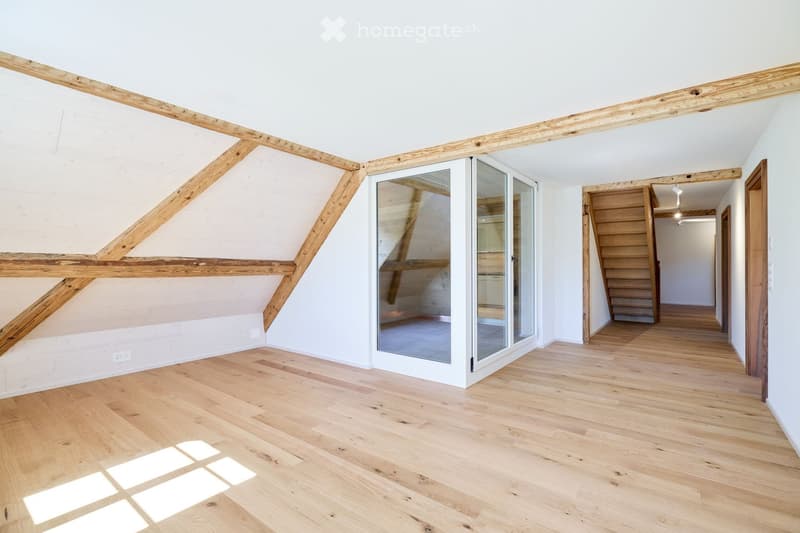 Bright, empty room with wooden flooring and exposed wooden beams. Features a large glass sliding door leading to another area and a wooden staircase in the background.