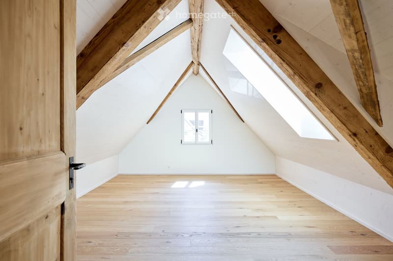 Bright, airy attic space with exposed wooden beams, white walls, and wooden flooring. Features a small window and a door leading to another area.