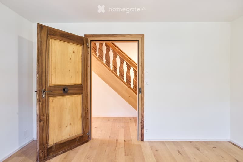 Empty room with light wooden flooring and white walls, featuring an open wooden door leading to a staircase with wooden railings.